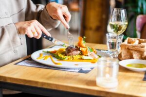 En person skär i en kötträtt på en restaurang med ett glas vitt vin bredvid.